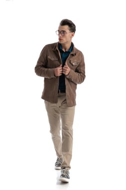 Öğrenci kahverengi deri ceketini ilikliyor ve beyaz arka planda ilerlerken yanına bakıyor.