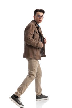 Yakışıklı, zeki, sıradan bir adam beyaz arka planda yürürken deri ceketli yakalı, tam vücut fotoğraflı.