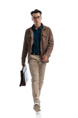 Genç iş adamı deri ceket giyiyor ve elinde evrak çantasıyla beyaz arka planda yürüyor.