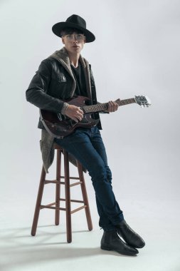 Şapkalı, gözlüklü, siyah deri ceketli, sandalyeye yaslanmış ve gri arka planda gitar çalan seksi adam.