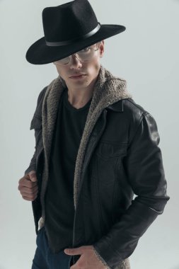 Şapkalı, gözlüklü, siyah deri ceketli ve gri arka planda poz veren çekici bir adam.