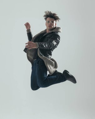 Siyah deri ceketli, gri arka planda havaya zıplayan dinamik havalı adamın yan görüntüsü.
