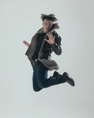 Havaya sıçrayan siyah deri ceketli havalı genç adam ve gri arka planda el hareketi yaparken dinamik olmak.