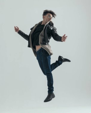 Siyah deri ceketli dinamik genç adam gri arka planda arkasına bakıyor ve havaya zıplıyor.