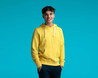 Sarı kapüşonlu rahat bir adam mavi arka planda durup gülümserken cebini tutuyor.