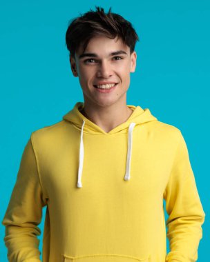 Mavi arka planda elleri ceplerinde dururken sarı kazak giyen çekici genç adamın portresi