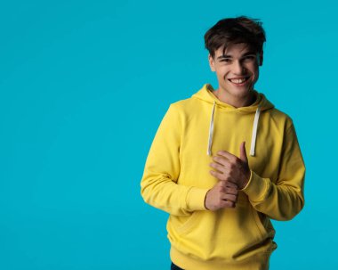 Mavi arka planda kollarını düzeltirken sarı kazak giyen mutlu ve çekici genç adamın portresi.