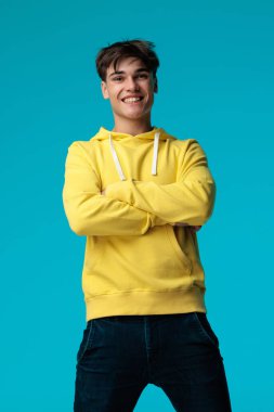 Kendine güvenen, sarı kapüşonlu, kollarını kavuşturmuş, gülümseyen, mavi arka planda bacaklarını açmış bir adamın yakın çekimi.