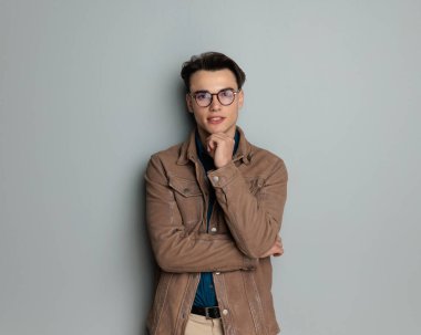 Gözlüklü, çenesine dokunan ve gri arka planda düşünen düşünceli bir gencin portresi.