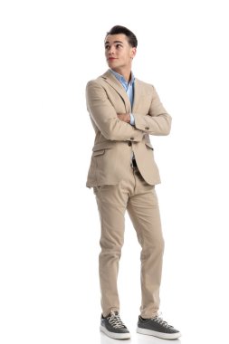 Kendine güvenen, bej takım elbiseli ve mavi gömlekli bir işadamı beyaz arka planda kolları bağlı dururken yukarı doğru bakıyor.