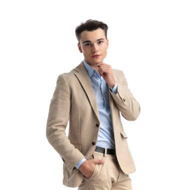 Bej takım elbiseli seksi öğrencinin beyaz arka planda dururken cebini ve çenesini tuttuğu yakın plan.