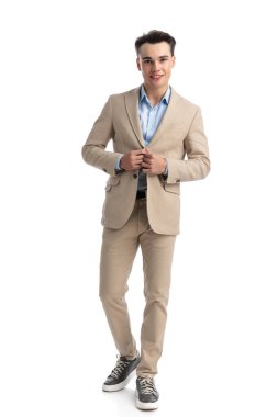 Seksi, zeki, sıradan bir adam bej takımının düğmelerini açıyor ve beyaz arka planda tek başına dururken gülümsüyor.