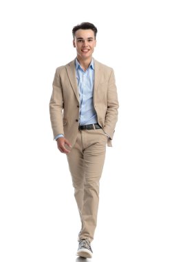 Beyaz arka planda yürürken cebinde cebi olan bej takım elbiseli mutlu ve rahat bir adam.