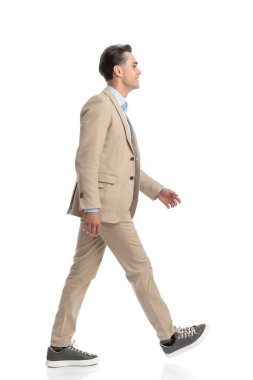 Beyaz arka planda yürüyen, bej takım elbiseli, mavi gömlekli, akıllı, sıradan bir adamın yan görüntüsü.