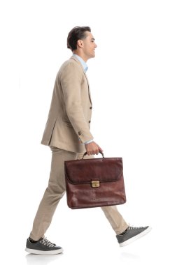 Beyaz arka planda yürüyen, bej takım elbiseli, evrak çantalı, sıradan bir gencin yan görüntüsü. 