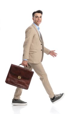 Bej takım elbise giyen ve elinde bavul tutan genç bir işletme öğrencisi kameraya bakarken beyaz arka planda yürüyordu.