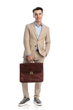 Bej takım elbiseli rahat iş adamı beyaz arka planda gülümserken cebini ve çantasını tutuyor.