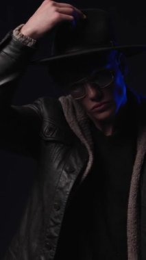 Gözlüklü ve siyah deri ceketli çekici genç adam şapkasını indiriyor ve siyah arka planda gizemli bir şekilde uzaklaşıyor.