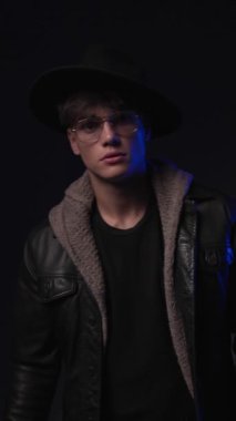 Siyah deri ceketli, gözlüklü, şapkasını çıkartıp siyah arka planda ilerleyen seksi bir genç.