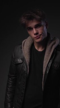 Siyah deri ceketli kibirli bir adam başını yana eğmiş gri arka planda ilerlerken ileriye bakıyor.