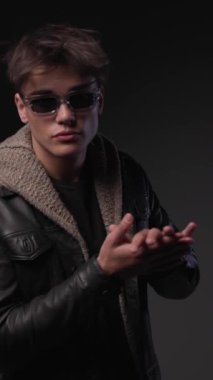 cool fashion man wearing transparent frame sunglasses, rubbing palms and fixing black leather jacket and glasses before walking away on grey background