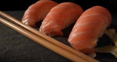 Pişmemiş pembe somon balığının, Nigiri suşisine dönüştürülmüş, gari ve wasabi ile servis edilen yumuşak pirinçli, ahşap çubuklu, siyah tabakta servis edilen videosunu kapat.