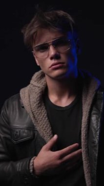 Siyah deri ceket açıp hareket eden, siyah arka plandaki kötü adam gibi davranan 20 'li yaşlarda kibirli bir gencin dikey videosu.