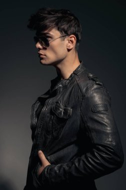 Siyah deri ceketli ve güneş gözlüklü genç motosikletçinin yan görünüşü elleri siyah arka planda duruyor.