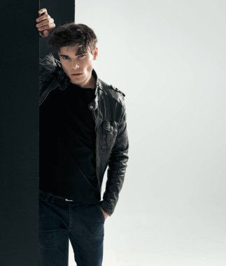 Gri arka planda siyah panele yaslanmış siyah deri ceketli rahat bir adamın yakın çekimi.