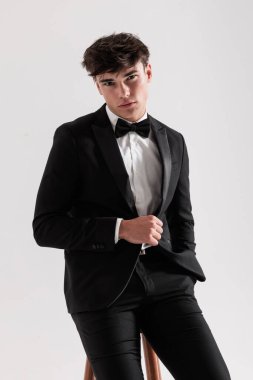 Siyah takım elbiseli ve papyonlu seksi moda adamı yakın plan. Gri arka planda tek başına otururken ahşap sandalyede oturuyor.