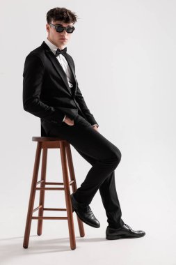 Siyah smokin ve güneş gözlüğü takan çekici işadamlarının yan görünüşü. Gri arka planda, tahta sandalyede otururken cepleri tutuyor.