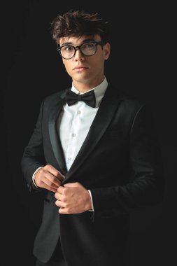 Siyah arka planda tek başına dikilirken siyah smokinini ilikleyen yuvarlak gözlüklü yakışıklı iş adamının yakın çekimi.