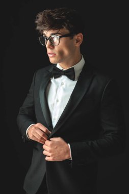 Gözlük takan yakışıklı damat siyah smokini ayarlıyor ve siyah arka planda dikilirken yan tarafa bakıyor.