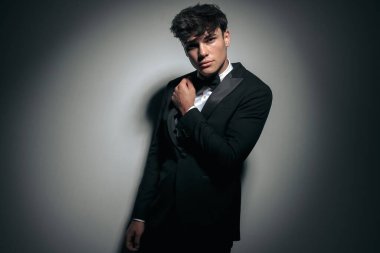 Siyah takım elbiseli ve gri duvar arkasında papyon tutan baştan çıkarıcı alfa erkek.