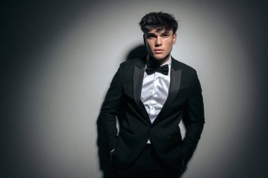 Siyah smokinli çekici bir delikanlının ceplerini tutarken koyu arkaplanda gri duvara yaslanırken.