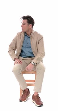 Her iki tarafa bakan, kollarını hareket ettiren, çenesine dokunan ve düşünen, gülümseyen, el hareketi yapan ve beyaz arka planda otururken ceketini tamir eden genç bir adam.