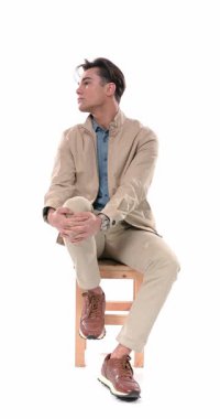 Elleriyle dizini tutan, yan bakan, bacak değiştiren, mutlu ve gülümseyen, beyaz arka planda oturan meraklı genç adam.