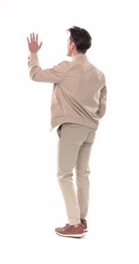 Bej ceketli sıradan bir genç adam beyaz arka planda kameranın önünde dikilirken el sallıyor.