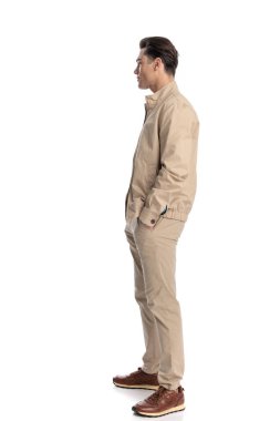 Bej ceketli, cebinde el ele tutuşan, beyaz arka planın önünde duran havalı genç adamın yan görüntüsü.