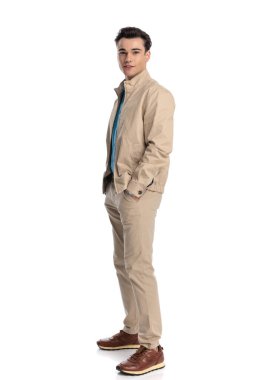 Mutlu genç bir adamın gülümseyen uzun bir resmi. Beyaz arka plan önünde elleri ceplerinde poz verirken.