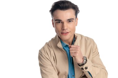 Bej ceketli sıradan bir genç öne bakıyor ve gülümsüyor. Stüdyoda beyaz arka planda izole bir şekilde poz veriyor.