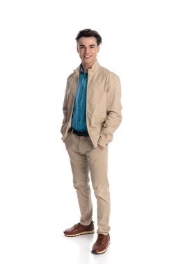 Sırıtan bej ceketli, çini pantolonlu, spor ayakkabılı, elleri ceplerinde beyaz arka planda mutlu bir adam.