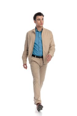 Uzun boylu, bej ceketli, Chino pantolonlu ve spor ayakkabılı, beyaz arka planda yürüyen çekici bir adamın resmi.