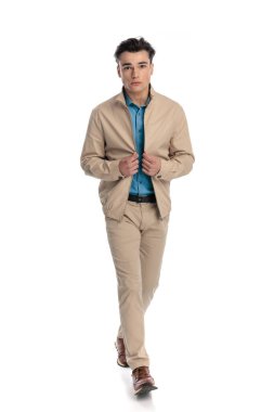 Yakışıklı genç adam ileriye bakıyor, ceketini kapatıyor ve beyaz arka planın önünde yürüyor.