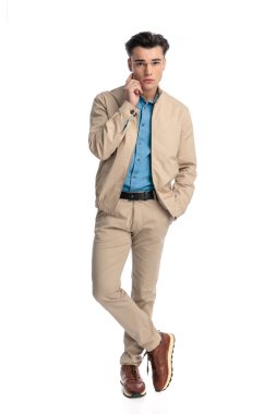 Bej ceketli, yanağa dokunan, elleri ceplerinde ve bacakları beyaz arka planda çapraz duran seksi bir genç adam.