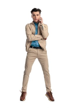 Bej ceketli, çenesine dokunan ve kollarını kavuşturup beyaz arka planda havada zıplayan mutlu genç adam.