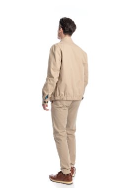 Beyaz arka planda yürüyen bej ceketli ve spor ayakkabılı sıradan bir genç adam.