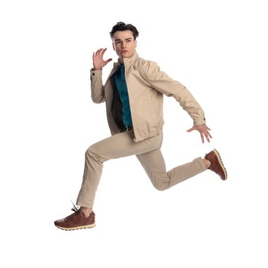 Dinamik genç adam, bej ceketli, Chino pantolonlu ve spor ayakkabılı büyük bir hamleyle havaya sıçrıyor ve beyaz arka plana bakıyor.