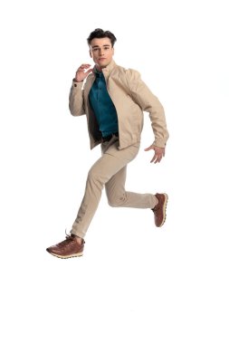 Beyaz arkaplan önünde poz verirken havada zıplayan bej ceketli sıradan dinamik adam.