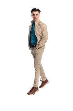 Kendinden emin, bej ceketli, elleri ceplerinde yürüyen ve beyaz arka planda ileriye bakan bir adam.